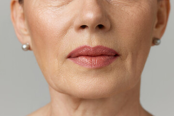 Cropped portrait of elderly woman with beautiful skin, plump lips over grey studio background. Mimic wrinkles.
