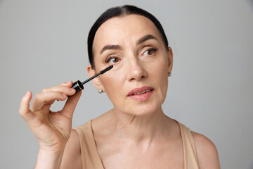 Close up portrait of beautiful mature brunette woman applying mascara with makeup brush over grey background. Fashion beauty.