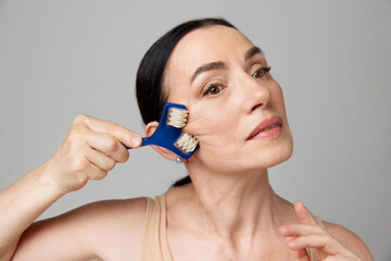 Mature woman using facial roller for smooth, anti aging and lifting treatment on a grey studio background.