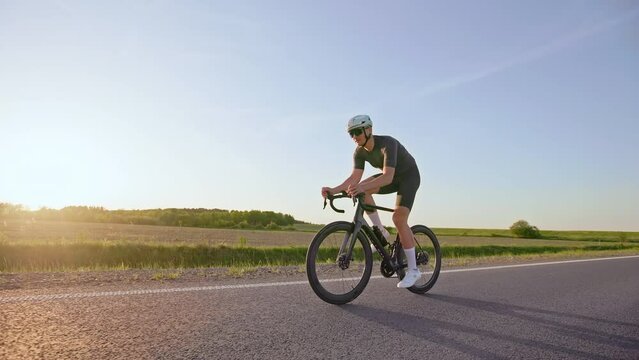 Full length view of athletic man on light road bike pedaling vigorously along paved highway at summer sunset. Experienced sports person training outside cycling at dynamic-intensity activity.