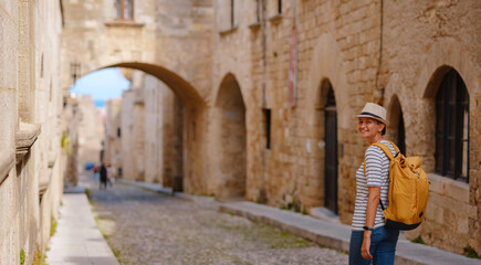 Fototapeta na wymiar summer trip to Rhodes island Greece. Young Asian woman in striped tshirt and hat walks Street of Knights of Fortifications castle. female traveler visiting southern Europe. Unesco world heritage site