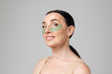 Portrait with mature woman using eye patches to get rid of eyes dark circles over grey background. Morning beauty routine
