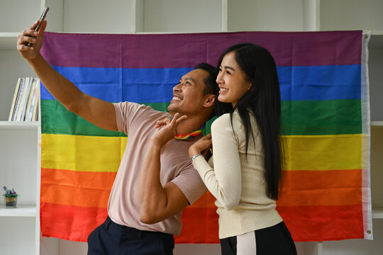 Cheerful biracial transgender man and young woman making selfie on smartphone against rainbow flag - Powered by Adobe