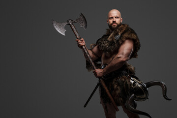 Fototapeta na wymiar A strong and intimidating Viking man in a beard and bald head, dressed in animal fur and light armor, with a helmet hanging from his belt, brandishing a huge axe against a gray background