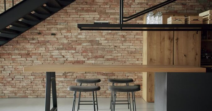 Modern and spacious wooden kitchen room with wall bricks and black staircase. Real Estate. Luxury kitchen with wooden island and chairs. Modern interior in luxury house. Minimalist design in New Home.