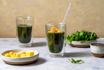 Green smoothie in glasses made with greens, frozen mango, and chia seeds, grey and yellow background, ingredients