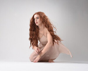 Fototapeta premium Full length portrait of beautiful brunette model wearing a pink dress. graceful sitting pose, kneeling on floor gestural hands. shot from low angle perspective, isolated on white studio background