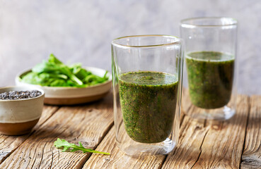 Green smoothie in two glasses on the wooden table and grey background, chia seeds, greens in ceramic bowl behind