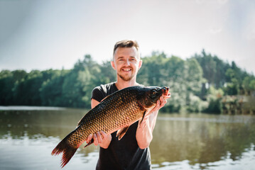 Happy fisherman hold big trophy fish carp near lake. Fresh fish trophy in hands. Young man returning with freshly caught fish. Article about fishing day. Fishing backgrounds. Success pike fishing.