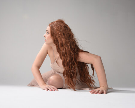 Full length portrait of beautiful brunette model  wearing a  pink dress. graceful sitting  pose, kneeling on floor gestural hands. shot from low angle perspective,  isolated on white studio background