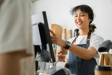 Happy young Asian woman cashier wears an apron and using pos terminal to input orders on coffee...