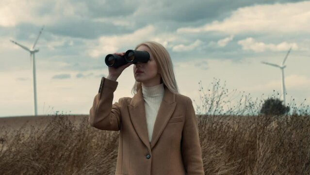 blonde woman in a stylish brown coat watching in binoculars in a dry grass meadow during autumn, Poland