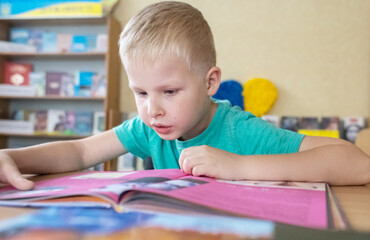 Boy in library. Seven year Ukrainian guy is sitting at a table in library and reading book. Concept...