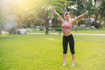 Happy fit female jogger. 30s Asian woman wearing pink sportswear breathing fresh air in public park. Wellness living and nature concept.