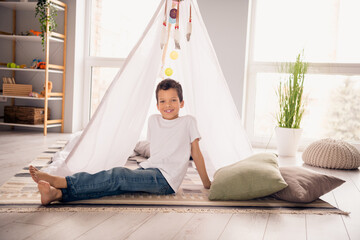 Portrait of adorable small schoolboy sitting floor build camping tent wear white shirt modern interior living room playroom daycare indoors © deagreez