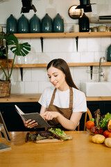 Young beautiful woman in the kitchen in an apron, fresh vegetables on the table, writes down her favorite recipes, comes up with ideas for dishes.