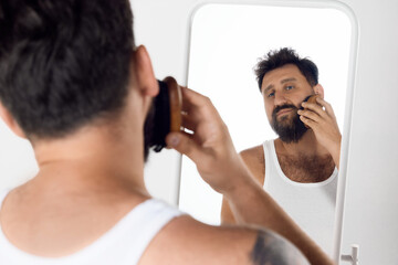 Mature man looking in mirror, taking care after beard, brushing with special brush over white studio background. Concept of men's beauty, skin care, cosmetology, health and wellness. Copy space for ad