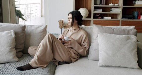 A beautiful Young woman is drinking coffee and tea in the living room at home, healthy calm lady relaxing on comfortable sofa feel stress free at home lounge alone. Woman rests at home on luxury sofa. - Powered by Adobe