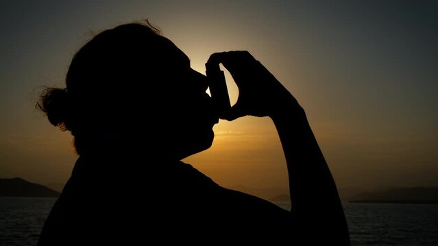 An asthma attack by the sea. Silhouette of a panting female using an inhaler in the evening on a sea beach.
