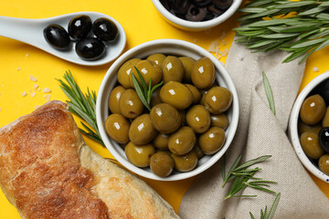 Olives in bowls and spoon, baguette on yellow background, top view
