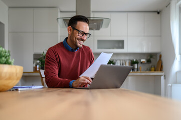 Happy male financial adviser reading income tax form and working over laptop on desk in home office