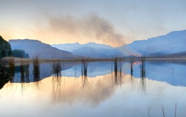 SMOKE FROM A WINTER GRASSFIRE reflects in a drakensberg mountain lake - 633338824