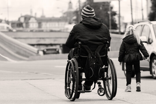 A disabled man rides in a wheelchair along the sidewalk, an elementary school daughter runs nearby, black and white photo. A man's legs were crippled after a spinal injury.