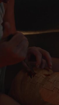  Vertical video. A young woman at night, surrounded by candles and covered in sweat, carves a face into a pumpkin for Halloween.