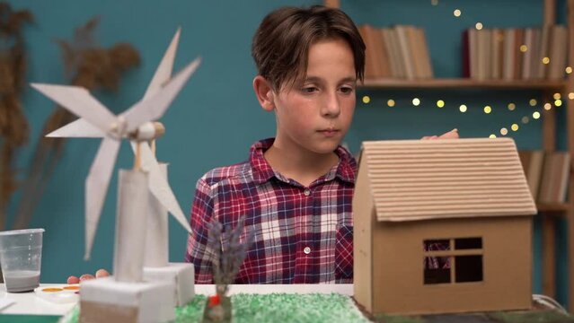 Attractive boy making creative project of wind turbines and cardboard house sitting at the table at home. Concept of scientific projects