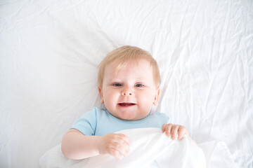 A baby with blonde hair smiling laying on a white bed. top view