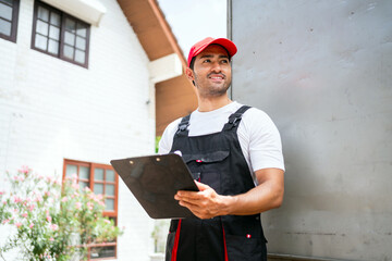 Asian truck driver wearing red cap holding a clipboard, checking the delivery packages checklists or paperwork and standing with a truck. Shipping cargo service, freight truck and transport logistics.