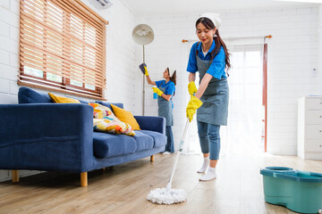 Two Asian young professional cleaning service women worker team working in the house. The girl housekeeper wiped a wet mop on wooden floor with another one clean the lamp with a feather duster.