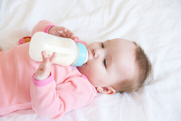 the baby drinks milk from a bottle lying on bed.