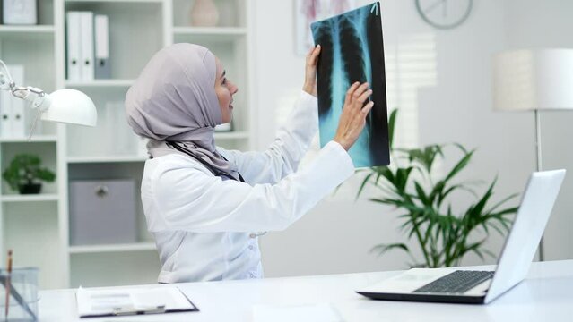 Confident muslim female doctor examining x-ray picture while sitting at workplace in office in hospital clinic. Woman in hijab, medical worker physician is carefully studying a picture of the lungs