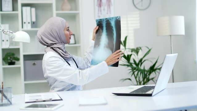 Muslim female doctor talking on a video call using a laptop while holding an x-ray in a modern hospital clinic. Medical worker woman physician consults a patient sitting at workplace in office