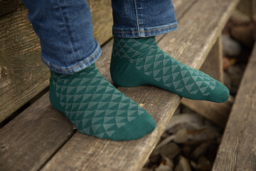 Outdoor product photo of men's teal patterned dress socks. The man is wearing jeans with them and is sitting on a wooden deck.