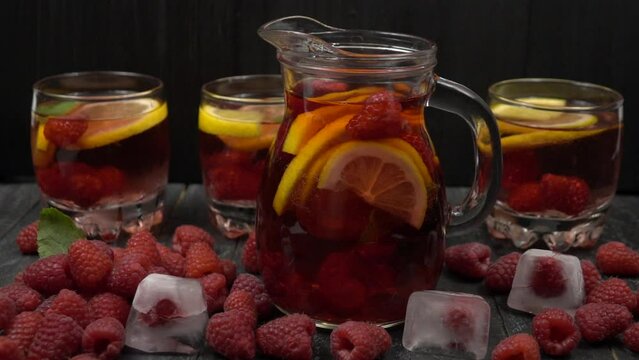 Ice cubes fall in a pitcher with homemade raspberry-citrus lemonade, slow motion.  Fresh raspberries, pitcher and glasses with lemonade are on dark wooden table, slow motion 