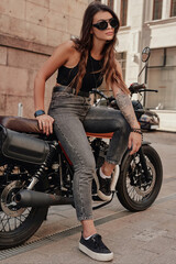 Stylish brunette in jeans and a tank top, with sunglasses and a tattoo on her arm, poses on her retro motorcycle on an old cobblestone street in Europe