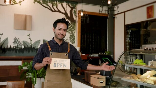 Indian man owns a coffee shop Smiling in overalls standing in the middle of a small coffee shop that is a family business In the hand there is a sign that says OPEN and points to glass cake cabinet.