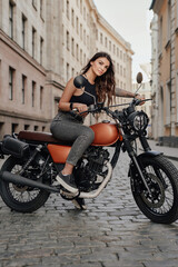 Obraz na płótnie Canvas In the old-world charm of a paved alleyway in Europe, a beautiful young woman with dark hair stands proudly next to her vintage motorcycle