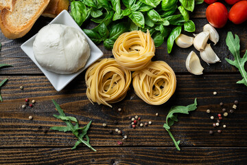 Italian food ingredients, fresh mozzarella, bruschetta, raw tagliatelle, garlic, tomatoes, arugula and basil leaves on a dark wooden background