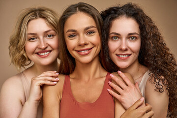 Close-up portrait of three beautiful different young women on a beige background who are smiling....