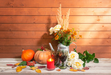 Bouquet of white roses, helenium and dry grass in watering can, candles, pumpkins, glasses of red wine and white roses near on aged wooden table.