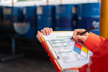 A safety officer is checking on the hazardous material checklist form with chemical storage area at the factory as background. Industrial safety working scene, selective focus. - obrazy, fototapety, plakaty