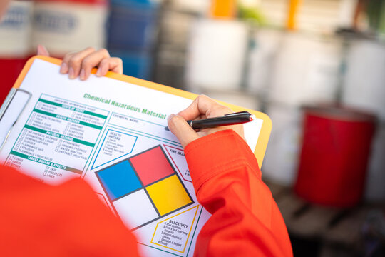 A safety officer is checking on the hazardous material checklist form with chemical storage area at the factory as background. Industrial safety working scene, selective focus.