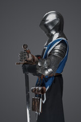 Medieval guard dressed in armor and blue surcoat holding a sword, standing in statue pose, against a gray background