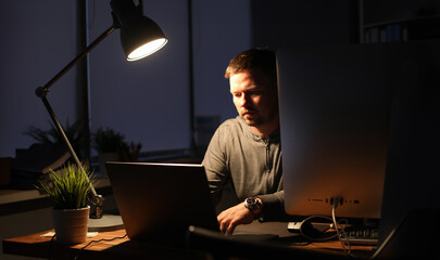 Businessman sits in an evenings office at table by light of lamp. Uses laptopand pc. The writer...
