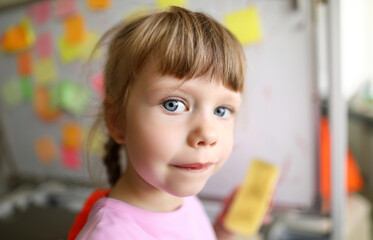 Focus on female child looking at camera with strange and unreadable glance on cute face and standing in modern classroom with high-tech glass board. Blurred background