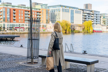 Blonde woman wearing a long beige coat and sunglasses walks after shopping with a paper bag and a bag in Grand Canal Square Dublin, Docklands, Dublin, Ireland