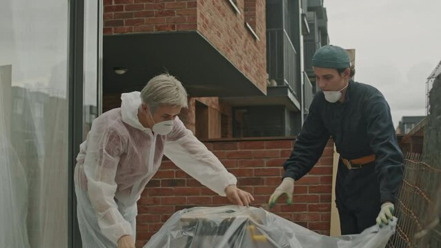 Medium shot of two young male construction workers covering portable workbench with polythene sheeting on balcony of renovating apartment building
